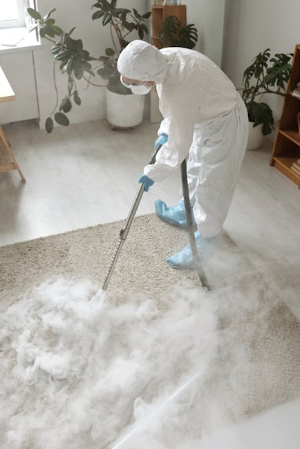 A professional carpet cleaner from Carpet Cleaners Kennington is performing deep surface cleaning in a residential living room. The technician is dressed in a full white protective suit, gloves, and a face mask, operating a professional carpet cleaning machine that is releasing steam into a beige, textured carpet. The room features a light-colored floor, with a window allowing natural light to illuminate the space. There are potted plants on a windowsill and a wooden shelf in the background, creating a clean, organized environment. The image emphasizes thorough sanitisation and deep cleaning processes aimed at maintaining hygiene and freshness in domestic spaces.
