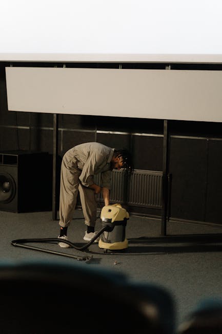 A professional cleaner from Carpet Cleaners Kennington is performing deep surface cleaning in a conference or event space. The individual, dressed in a light beige coverall, is bent over, using a vacuum or extraction machine to thoroughly clean the carpeted floor. The room features dark paneling on the walls, and a large white projection screen or panel is mounted above, illuminated by overhead lighting. The space appears tidy and well-maintained, with visible cleaning equipment suggesting a focus on hygiene and deep cleaning processes to ensure a sanitized environment. The scene reflects a professional approach to commercial cleaning services, emphasizing surface cleaning and maintenance in a corporate setting.