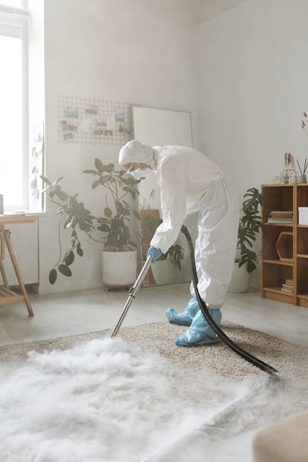 A professional carpet cleaner from Carpet Cleaners Kennington is performing deep surface cleaning in a residential living room. The technician is dressed in a full white protective suit, gloves, and a face mask, operating a professional carpet cleaning machine that is releasing steam into a beige, textured carpet. The room features a light-colored floor, with a window allowing natural light to illuminate the space. There are potted plants on a windowsill and a wooden shelf in the background, creating a clean, organized environment. The image emphasizes thorough sanitisation and deep cleaning processes aimed at maintaining hygiene and freshness in domestic spaces.