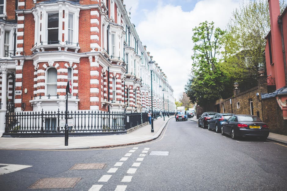 A street view of Kennington with Victorian-style terraced houses featuring red brick facades, white decorative window frames, and black metal fencing along the sidewalk. The street includes parked cars on the right side, modern vehicles in dark shades, and a light-colored bus in the distance. The road is paved with asphalt and marked with white dashed lines, while the sidewalk is concrete with tactile paving. Trees with fresh green foliage line the right side, providing shade and contrast against the buildings. Bright daylight and a partly cloudy sky illuminate the scene, highlighting the clean and well-maintained appearance of the urban environment. Carpet Cleaners Kennington specializes in surface cleaning and deep cleaning services that ensure hygiene and upkeep of residential and commercial spaces across this area.