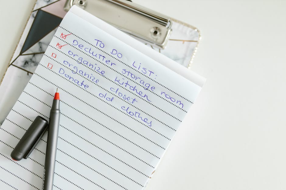 A close-up of a handwritten to-do list on a spiral-bound notepad placed on a white surface, with the list titled 'TO DO LIST' written in blue ink. The tasks include 'Declutter storage room', 'Organise kitchen', 'Organize closet', and 'Donate old clothes', each marked with a red checkmark beside the first two tasks. A silver and orange ballpoint pen with its cap removed rests diagonally across the notepad, adjacent to a small metallic folder or binder in the background. The scene is well-lit, emphasizing a clean and organized workspace related to domestic surface cleaning and home organisation, as part of a typical end of tenancy cleaning checklist by Carpet Cleaners Kennington.