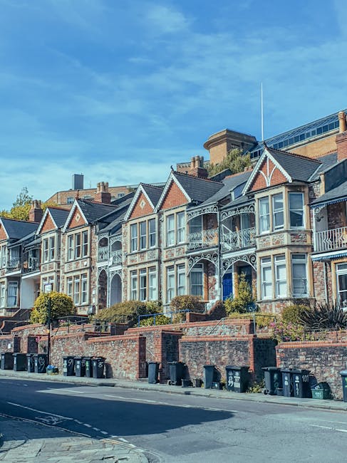 A street view of Kennington with Victorian-style terraced houses featuring red brick facades, white decorative window frames, and black metal fencing along the sidewalk. The street includes parked cars on the right side, modern vehicles in dark shades, and a light-colored bus in the distance. The road is paved with asphalt and marked with white dashed lines, while the sidewalk is concrete with tactile paving. Trees with fresh green foliage line the right side, providing shade and contrast against the buildings. Bright daylight and a partly cloudy sky illuminate the scene, highlighting the clean and well-maintained appearance of the urban environment. Carpet Cleaners Kennington specializes in surface cleaning and deep cleaning services that ensure hygiene and upkeep of residential and commercial spaces across this area.