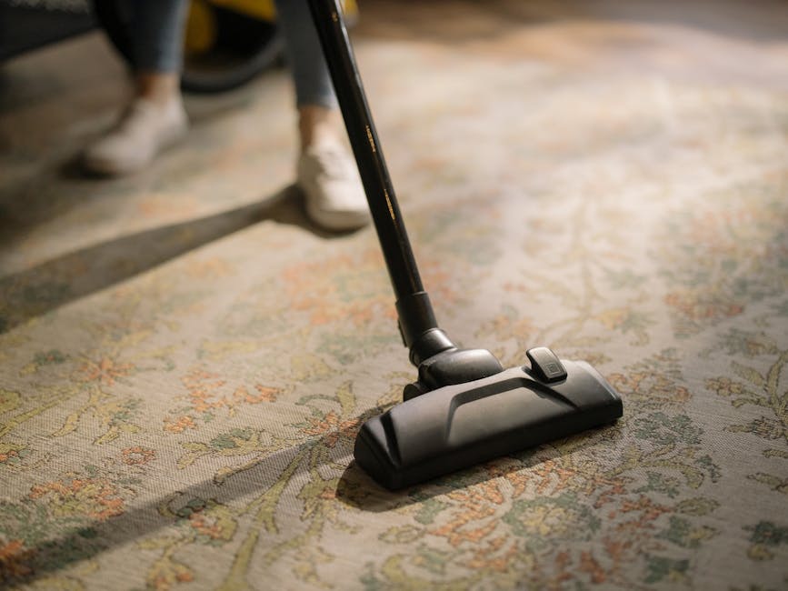Close-up of a black vacuum cleaner head actively cleaning a patterned, beige and green area rug in a well-lit room, with a person's legs and sneakers partially visible in the background. The surface of the carpet appears clean and free of visible dust or debris, demonstrating a deep cleaning process. The setting suggests a domestic environment, with natural light enhancing the cleanliness of the space, consistently reflecting professional surface cleaning services by Carpet Cleaners Kennington, as described on the Kennington Road carpet cleaning services near The Oval page.