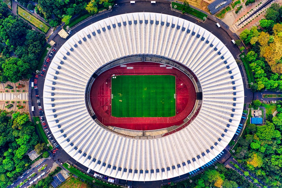 Aerial view of the Oval stadium in Kennington, SE11, showcasing an oval-shaped structure with a white, ribbed exterior roof surrounding the central pitch. The pitch is covered with vibrant green artificial turf marked with white lines, encircled by a red running track. Adjacent areas include landscaped greenery, trees, pathways, and parking spaces, all under bright natural lighting, highlighting the stadium's clean and well-maintained appearance, consistent with surface cleaning and maintenance practices by Carpet Cleaners Kennington for optimal hygiene and presentation.