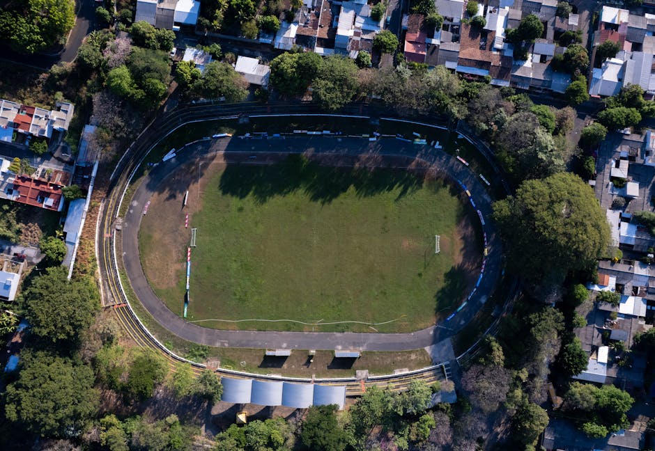 An aerial view of a sports stadium with a grass field in the center, surrounded by a running track and seating areas, located within a residential neighborhood with numerous trees and houses. The stadium appears clean and well-maintained, with the grass surface evenly green and free of debris. The surrounding pathways and seating areas are clear and tidy, reflecting routine surface cleaning and maintenance. The lighting suggests natural daylight, highlighting the neat and orderly appearance of the entire site. This image illustrates a clean, hygienic sports facility, consistent with professional deep cleaning and sanitisation practices by Carpet Cleaners Kennington, who specialize in surface cleaning and hygiene maintenance for various settings.