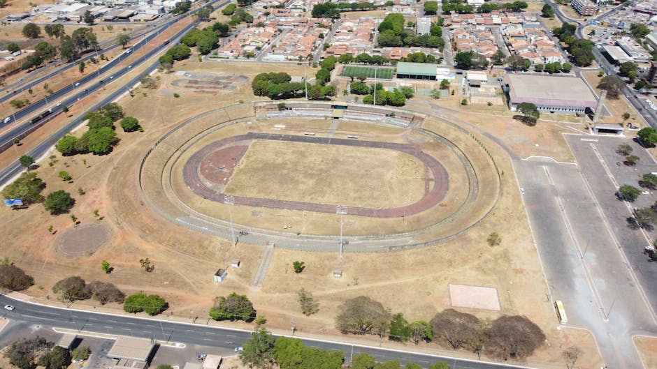 Aerial view of the Oval stadium in Kennington, SE11, showcasing an oval-shaped structure with a white, ribbed exterior roof surrounding the central pitch. The pitch is covered with vibrant green artificial turf marked with white lines, encircled by a red running track. Adjacent areas include landscaped greenery, trees, pathways, and parking spaces, all under bright natural lighting, highlighting the stadium's clean and well-maintained appearance, consistent with surface cleaning and maintenance practices by Carpet Cleaners Kennington for optimal hygiene and presentation.