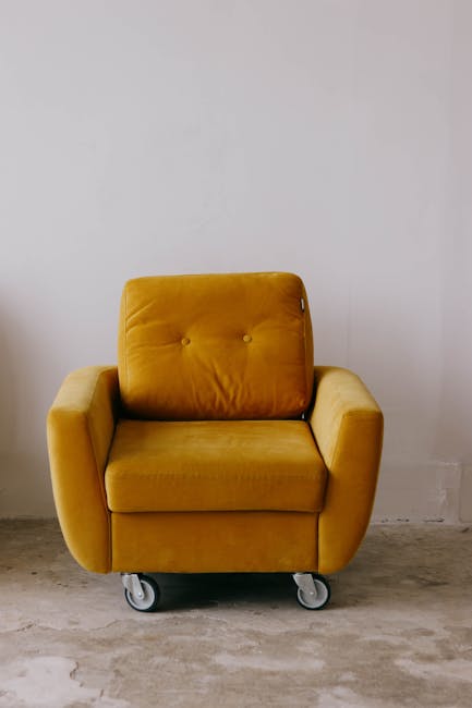 A mustard yellow upholstered armchair with a tufted backrest and seat cushion, featuring fabric material and slightly rounded armrests, positioned on four small caster wheels for mobility, set against a plain white wall with a concrete floor. The chair appears clean with no visible dust or stains, highlighting a neat and tidy appearance. The scene suggests a minimalist interior space, and the image is relevant to domestic or commercial surface cleaning, deep cleaning, or sanitisation services provided by Carpet Cleaners Kennington, as seen on their website for the SE11 upholstery cleaning guide for Kennington homes.