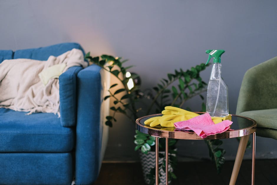 A living room corner featuring a blue upholstered sofa with a cream-colored throw draped over one arm and a pillow. Next to the sofa, there is a small round side table with a reflective surface, holding yellow rubber cleaning gloves, a pink cloth, and a transparent spray bottle with a green nozzle. Behind the table, there is a green upholstered armchair. In the background, a potted plant with broad green leaves adds a touch of nature. The room appears tidy and well-lit, emphasizing cleanliness and preparation for surface cleaning or sanitisation, aligned with the services offered by Carpet Cleaners Kennington for upholstery deep cleaning and domestic maintenance.